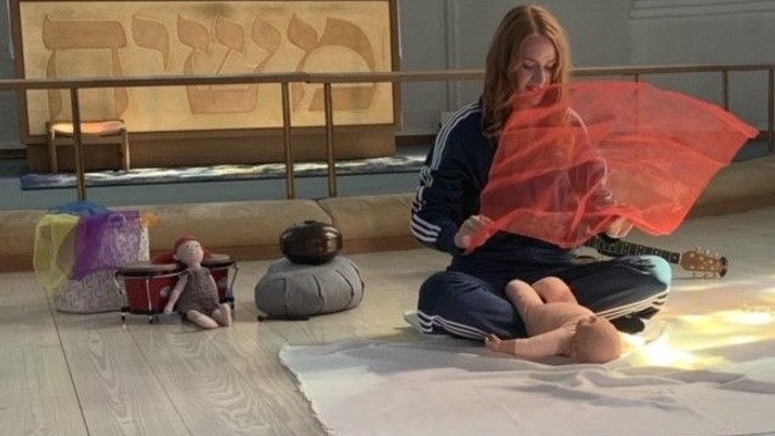 En kvinde med rød skjorte sidder på gulvmatte i en kirke med en rød paraply og diverse genstande omkring hende.