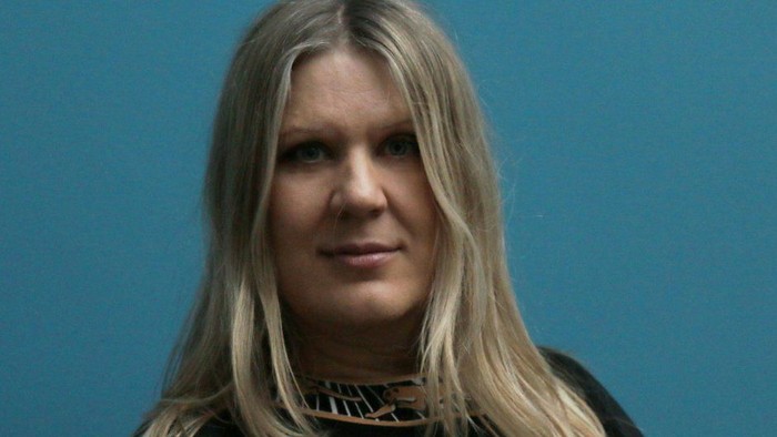 En kvinde med blond hår og mørkt tøj står foran et blåt baggrund.