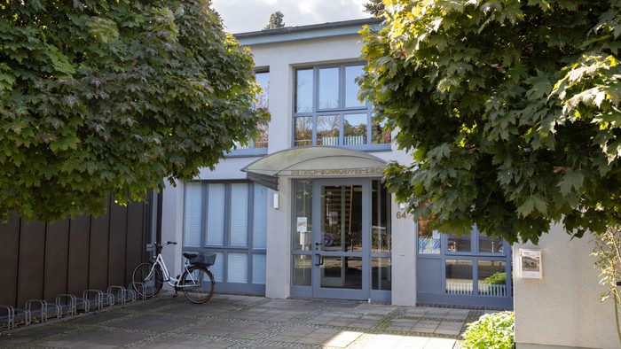 Das Bild zeigt ein weißes Gebäude mit Säulen und einem Vordach, umgeben von Bäumen und einem Zaun.