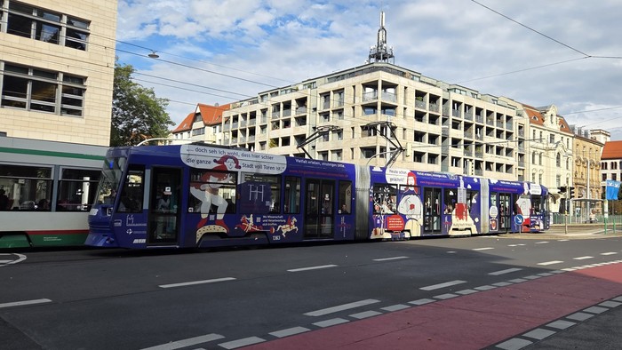 Moderne Straßenbahnen und Busse fahren an einer städtischen Kreuzung mit mehrstöckigen Gebäuden im Hintergrund.