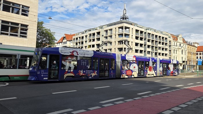 Moderne Straßenbahnen und Busse fahren an einer städtischen Kreuzung mit mehrstöckigen Gebäuden im Hintergrund.