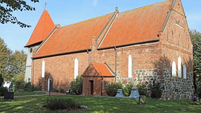 Das Bild zeigt eine rote Backsteinkirche mit spitzen Dächern und weißen Fenstern, umgeben von grünem Gras und Bäumen.