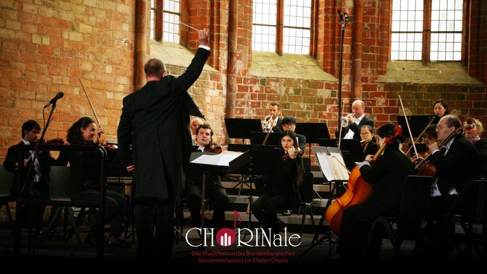 Ein Orchester probt unter Leitung eines Dirigenten in einem Raum mit Backsteinwänden und hohen Fenstern.