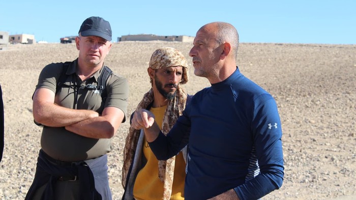 Tre mænd i udendørs omgivelser, hvor den ene peger, mens de andre observerer et fjernt område.