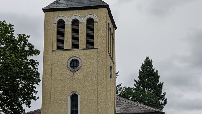 Kirchturm mit Kreuz und Uhr, gelb gestrichen, vor bewölktem Himmel