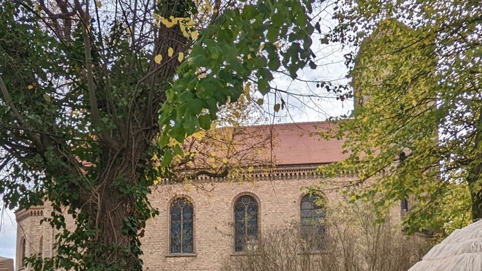 Altes Backsteingebäude mit rotem Dach und Uhrturm, umgeben von Bäumen und Büschen