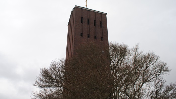 Ein hoher Turm ragt hinter Bäumen empor, der mit einem Kreuz gekrönt ist.