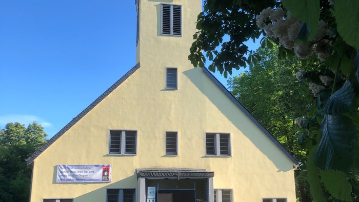 Gelbe Kirche mit Turmspitze und Uhr, Banner über dem Eingang hängend, umgeben von grünen Bäumen und Gras.