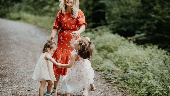 Eine Frau in einem roten Kleid geht Hand in Hand mit zwei jungen Mädchen in weißen Kleidern auf einem Feldweg.