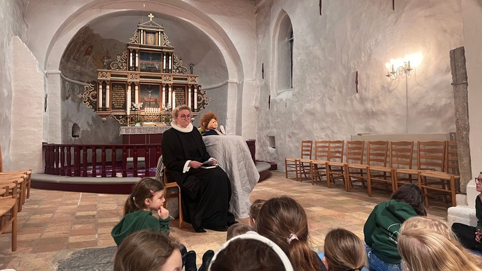 En nonne leder en gruppe børn i en gudstjeneste eller undervisning, hvor de sidder opmærksomt på træbænke.