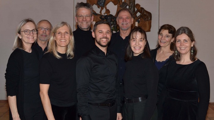 En gruppe på ni personer, der poserer sammen i formelt sort tøj foran en dekorativ baggrundsvæg.