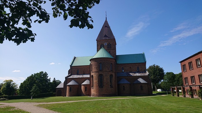 Historisk murstenskirke med grønt tag og højt spir, omgivet af træer og græs.