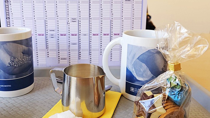 Ein Schreibtisch mit Kaffeetassen, Milchkännchen, Zuckerstücken und einem Korb mit Süßigkeiten vor einem Kalender.