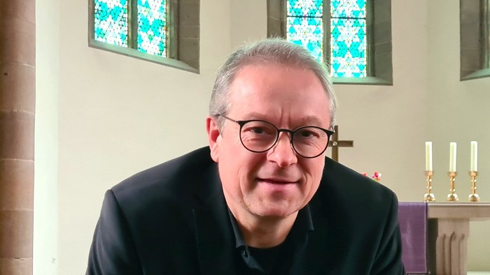 Mann mit Brille und dunklem Anzug, der in einer Kirche sitzt.