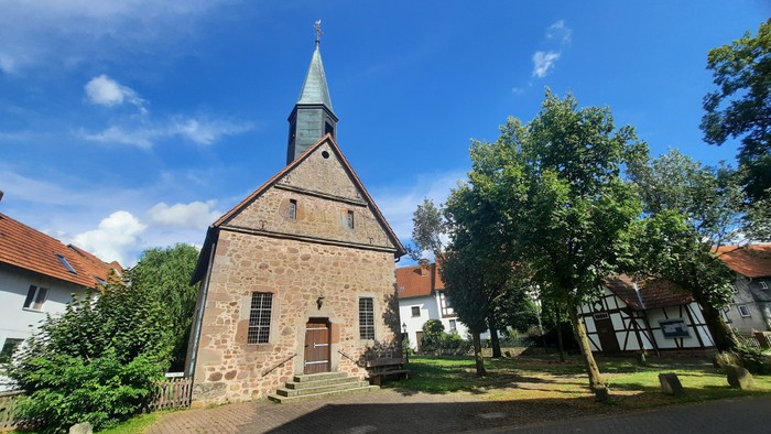 Kirchengrundstück mit altem Steingebäude und spitzem Dach, umgeben von Bäumen und anderen Häusern