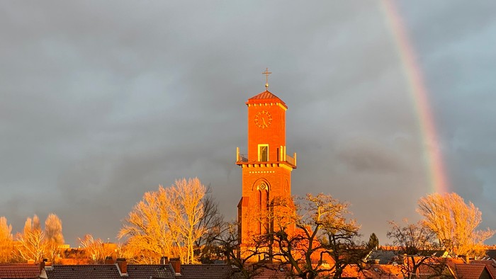 Eine auffällige orangefarbene Kirchturmspitze hebt sich vor einem dramatischen Himmel mit einem lebendigen Regenbogen ab.