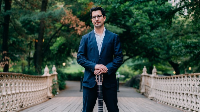 Ein Mann im Anzug hält eine akustische Gitarre auf einer Holzbrücke, umgeben von Grün.