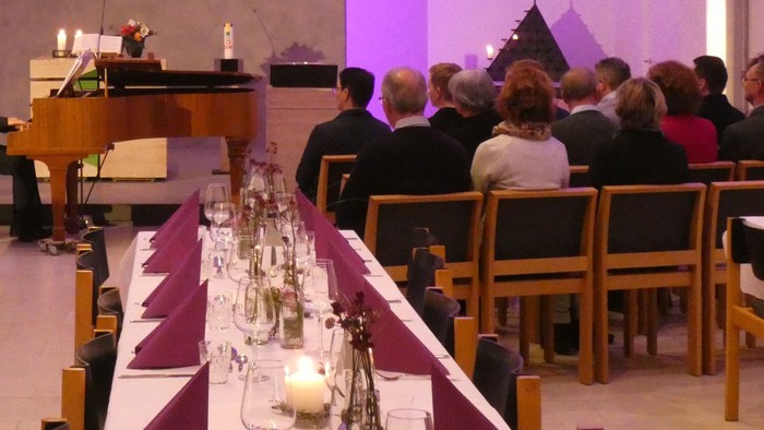 Eine feierliche Kirchenversammlung zeigt einen langen Tisch, der mit Kerzen gedeckt ist, flankiert von Teilnehmern und einem Pianisten am Altar.