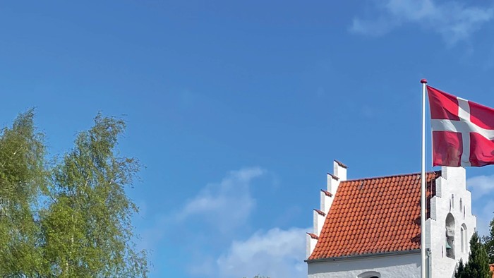 En hvid klokketårn med rødt tag og en dansk flag på toppen mod en klar blå himmel