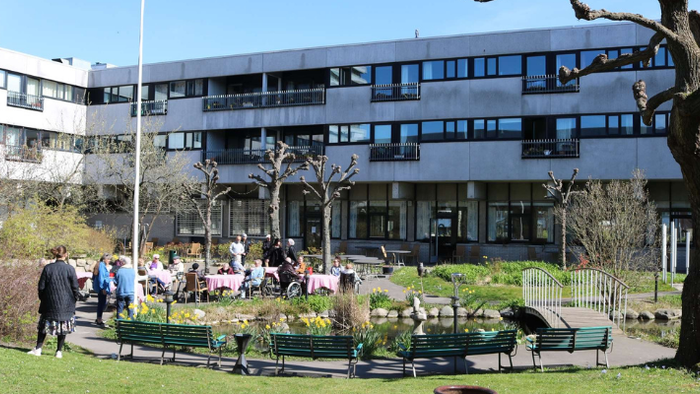 En gruppe af mennesker sidder på bænke i en park foran et stort bygning.