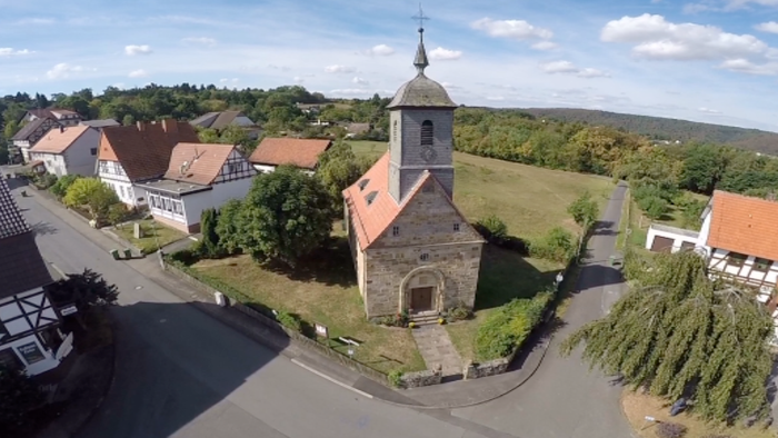 Luftaufnahme eines malerischen Dorfes mit einer kleinen Steinkirche, umgeben von Häusern und grünen Feldern.