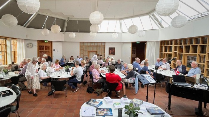 En gruppe ældre samles i et lys og rummeligt rum med hængende lyskugle og musikinstrumenter.