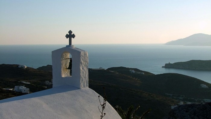 Weißer Kirchturm mit Kreuz vor Meer und Bergen