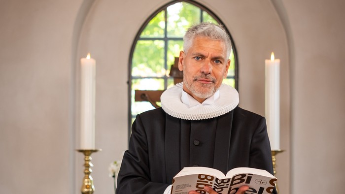 Mand i sort kåbe, der holder en bog i en kirke.