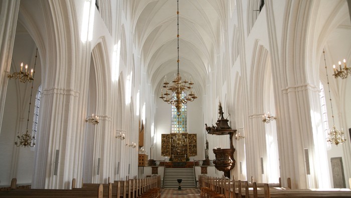 Indre del af en stor, belyset kirke med høje buegange og træbænke.