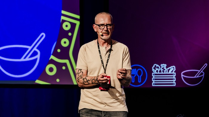 En taler på scenen, der diskuterer mad og ernæring, holder en rød genstand, mens vedkommende henvender sig til publikum.