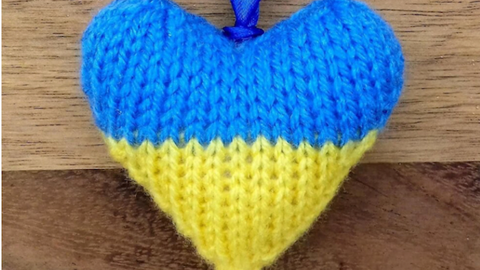 Gestricktes herzförmiges Ornament in Blau und Gelb, das an einer Holzfläche hängt.