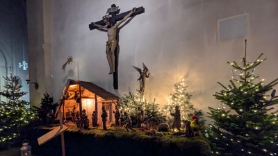 Aufwendige Weihnachtskrippe mit einem Kruzifix im Hintergrund, umgeben von geschmückten Weihnachtsbäumen und Kerzenlicht.