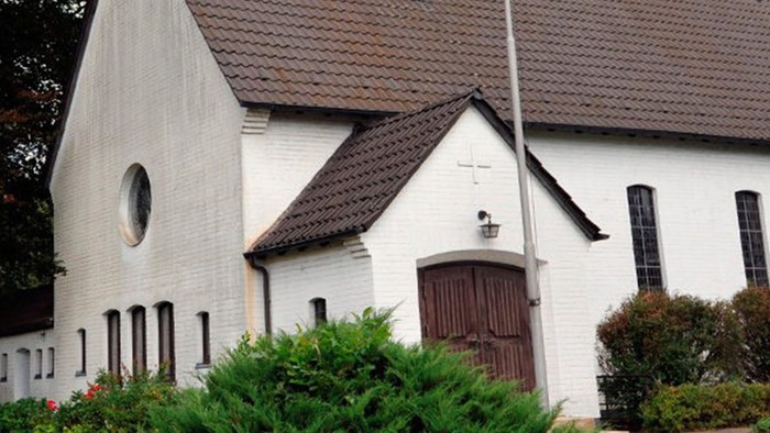 Weiße Kirche mit hohem Turm und braunem Dach, umgeben von Bäumen und Büschen