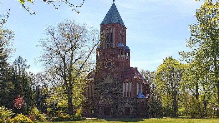 Das Bild zeigt eine rote Backsteinkirche mit hohem Turm in einem grünen Park.