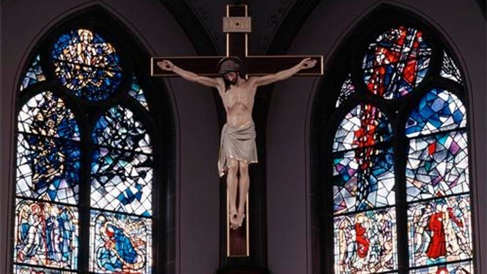 Das Bild zeigt ein Kruzifix vor zwei bunt gefärbten Kirchenfenstern.