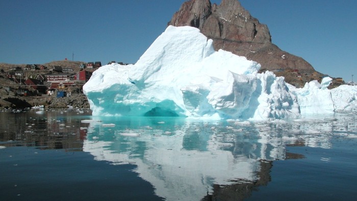 Kæmpe isbjerg flyder i stille vand ved siden af en klippefyldt kyst med en lille kystnær bebyggelse.