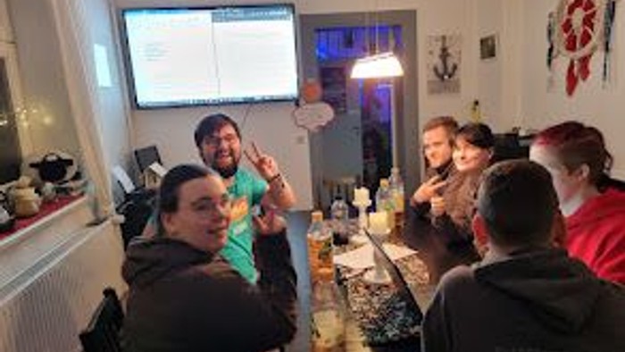 Eine Gruppe von Menschen steht um einen Tisch mit Getränken herum und schaut auf einen Fernseher.