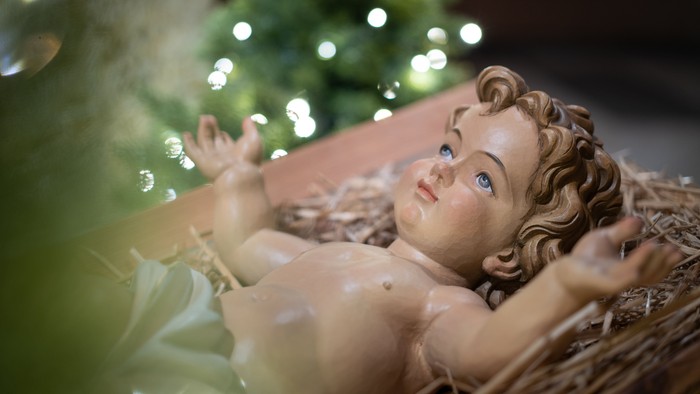Ein Babyfigur liegt in einem Korb, im Hintergrund ein beleuchteter Weihnachtsbaum.