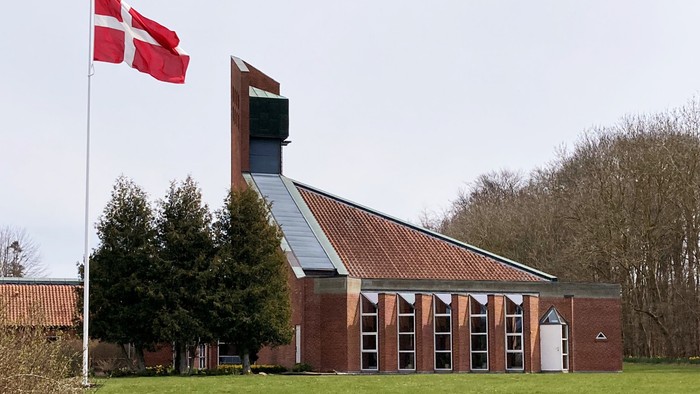 Moderne murstenskirke med det danske flag vajende, omgivet af grønt græsplæne og træer.