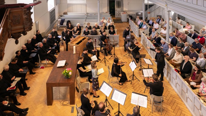 Ein Orchester probt in einem großen Saal, während Zuschauer zuschauen