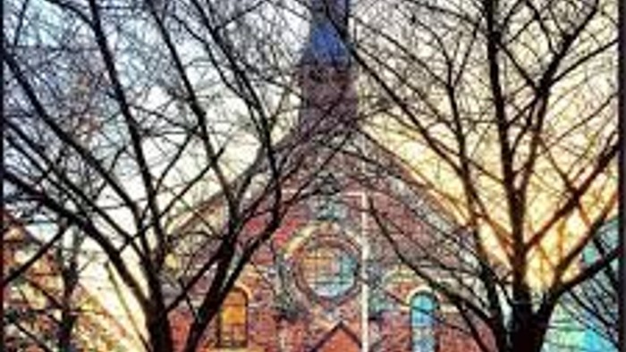 En rød murstenskirke med spiret mellem to store, blærede træer.