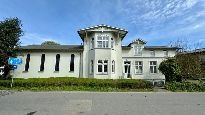 Ein großes, weißes Haus mit zahlreichen Fenstern und einem Tor im Vorgarten.