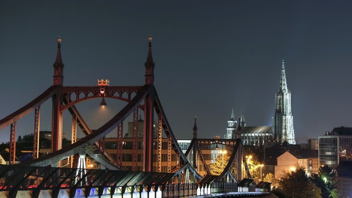 Ein beleuchteter roter Brückenbogen vor einer Stadtlandschaft bei Nacht