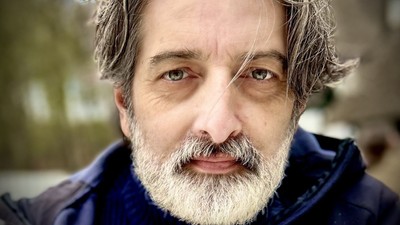 En mand med langt gråt hår og skæg iført en blå jakke udendørs.