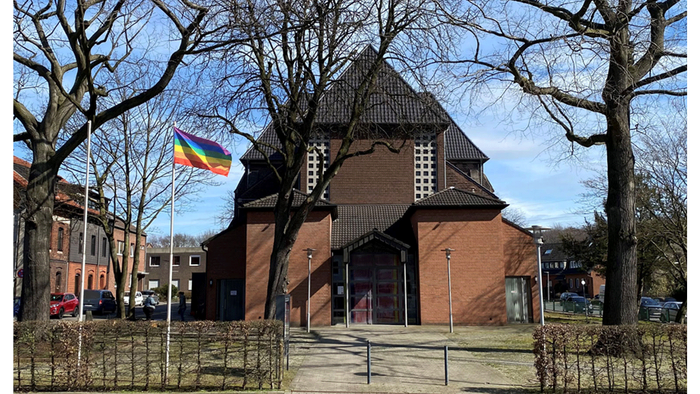 Historische Backsteinkirche mit einer Regenbogenflagge, die prominent davor weht, umgeben von kahlen Bäumen.