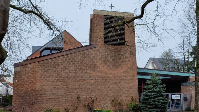 Ein Ziegelgebäude mit Uhrturm und Kreuz, umgeben von Bäumen und Büschen