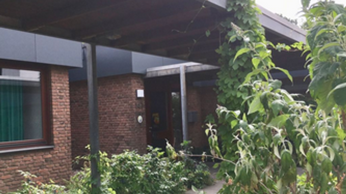 rotes Backsteinhaus mit Flachdach und überdachtem Weg zum Eingang, rechts und links davon begrünt