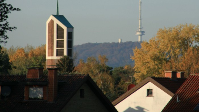 Städtische Skyline mit Kirchturm, Wohnhaustdächern und einem hohen Sendemast vor herbstlichem Laub.