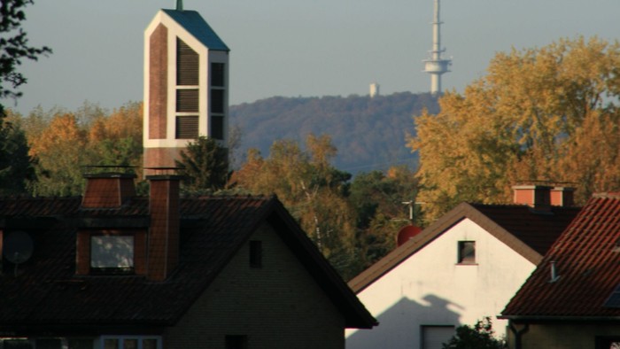 Städtische Skyline mit Kirchturm, Wohnhaustdächern und einem hohen Sendemast vor herbstlichem Laub.