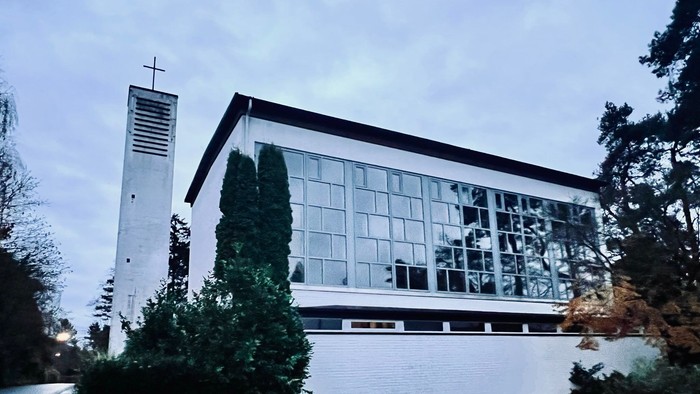 Moderne Kirchengebäude mit großen Glasfenstern und einem hohen Glockenturm neben üppigem Grün.